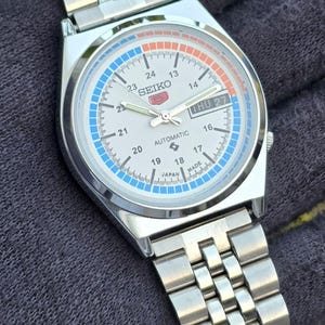 May include: A silver-toned Seiko automatic wristwatch with a white face, featuring blue and orange accents. The watch displays the day and date, with a stainless steel band. The watch face includes the text "SEIKO" and "AUTOMATIC".