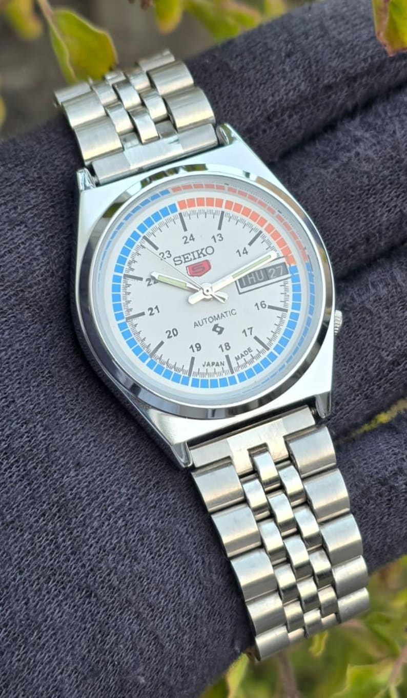 May include: A silver-toned Seiko automatic wristwatch with a white face, featuring blue and orange accents. The watch displays the day and date, with a stainless steel band. The watch face includes the text "SEIKO" and "AUTOMATIC".