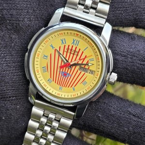 Pode incluir: Um relógio de pulso automático Citizen vintage com mostrador amarelo com detalhes em vermelho e azul. O relógio tem uma pulseira e caixa de aço inoxidável, com algarismos romanos e um visor de dia. O mostrador do relógio tem o texto "Citizen Automatic".