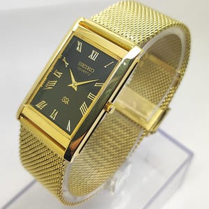 Könnte beinhalten: Eine goldfarbene Seiko Quartz-Uhr mit einem rechteckigen, dunkelgrünen Zifferblatt und goldenen römischen Ziffern. Die Uhr hat ein Mesh-Armband und ein goldfarbenes Gehäuse. Das Zifferblatt zeigt das Seiko-Logo und das Wort "Quartz".