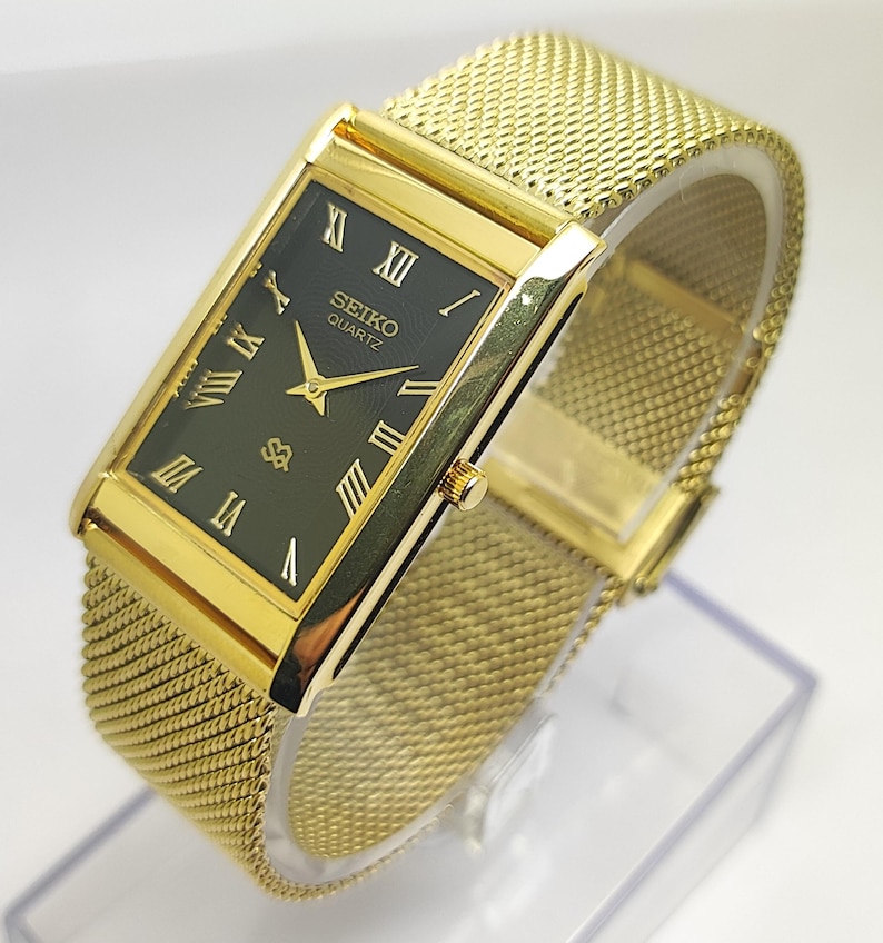 Peut inclure: Une montre Seiko Quartz dor&eacute;e avec un cadran rectangulaire vert fonc&eacute; et des chiffres romains dor&eacute;s. La montre a un bracelet en maille et un bo&icirc;tier dor&eacute;. Le cadran affiche le logo Seiko et le mot "Quartz".
