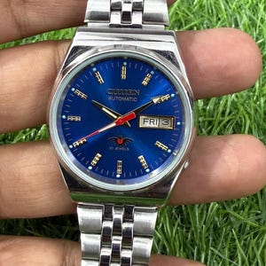 May include: A silver-toned Citizen automatic watch with a blue dial. The watch face displays the day and date, with the text "Citizen Automatic" and "21 Jewels". The watch has a silver link band.