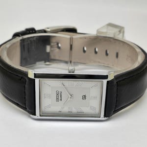 Puede incluir: Un reloj Seiko Quartz rectangular con esfera plateada y correa de cuero negro. La esfera del reloj presenta números romanos y el logotipo de Seiko. La caja es plateada y la correa de cuero negro.