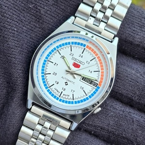 May include: Silver-toned Seiko automatic wristwatch with a white face, featuring blue and orange accents. The watch displays the day and date, with a stainless steel band. The face includes the text "SEIKO" and "AUTOMATIC".
