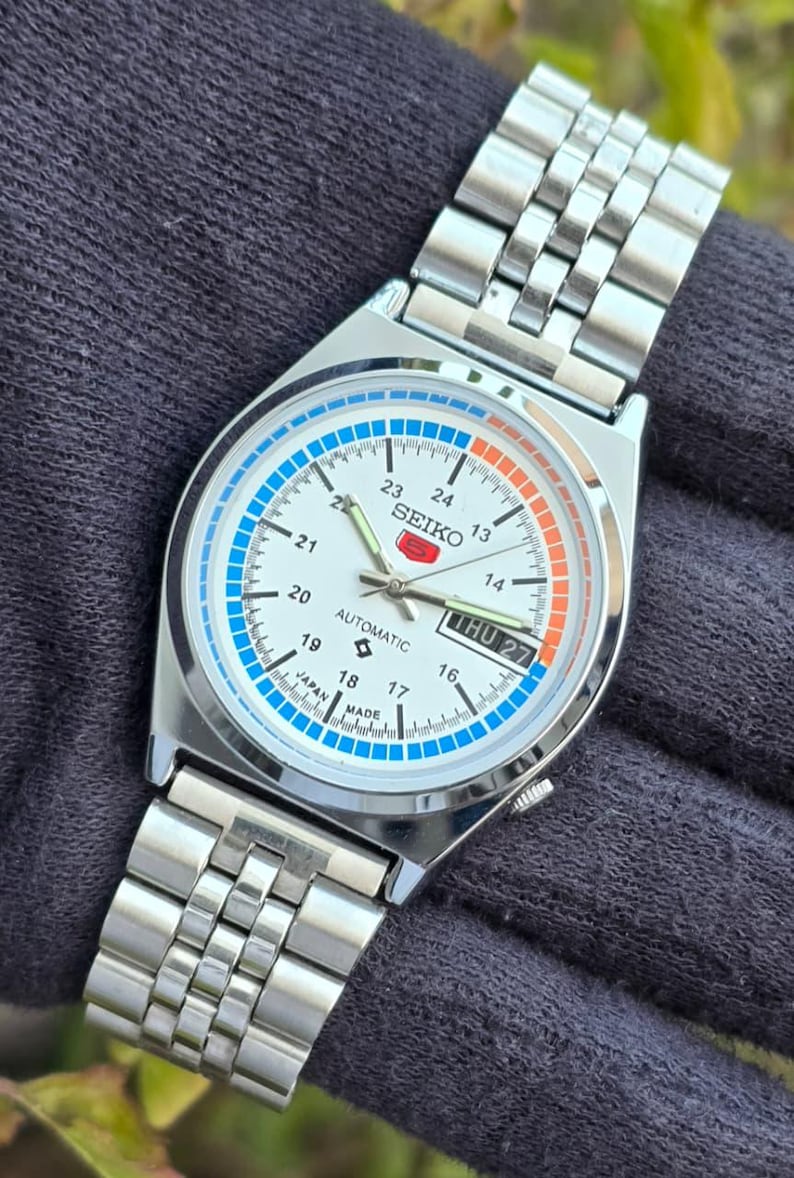 May include: Silver-toned Seiko automatic wristwatch with a white face, featuring blue and orange accents. The watch displays the day and date, with a stainless steel band. The face includes the text "SEIKO" and "AUTOMATIC".