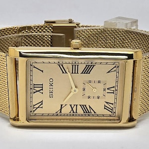 May include: A gold-toned Seiko wristwatch with a rectangular face and mesh band. The watch face features black Roman numerals and a smaller subdial. The watch is a classic design, suitable for formal occasions.