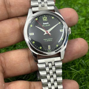 Puede incluir: Un reloj de pulsera plateado con esfera negra. El reloj muestra el texto "hmt Pilot" y "PARA SHOCK 17 JEWELS". El reloj tiene una correa plateada y está sujeto por una mano.