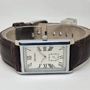 May include: A rectangular Seiko watch with a silver case and a cream-colored face featuring Roman numerals. The watch has a brown leather band. The watch face displays the Seiko logo and two smaller dials. The watch is a classic design.