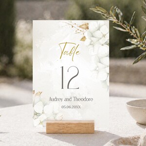 May include: A wedding table number card with the number 12 in black, and the word "Table" in gold script. The card features white floral and gold accents, with the names "Audrey and Theodore" and the date 05.06.20XX. It is displayed on a wooden block.