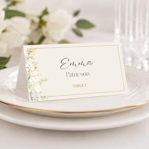 Może przedstawiać: Złożona wizytówka z nadrukowanym imieniem "Emma Patricson" i "Table 1", spoczywająca na białym talerzu ze złotą obwódką. Kwiatowy wzór akwarelowy po lewej stronie. W tle szklanka i kwiaty.