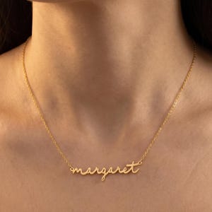 Può includere: Una collana color oro con il nome "margaret" in corsivo. La collana è indossata attorno al collo di una persona, con la catena e il ciondolo ben visibili. Il ciondolo è delicato ed elegante.