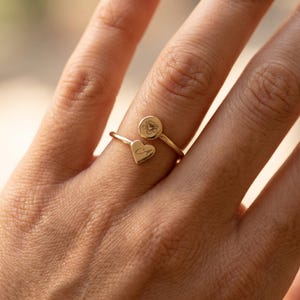 May include: A gold-colored ring with a heart and a circular charm. The heart charm has an engraved letter "A", and the circular charm features a floral design. The ring is worn on a finger.