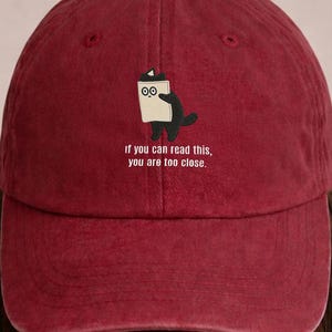 Könnte beinhalten: Burgunderfarbene Baseballkappe mit gebogenem Schirm. Die Kappe zeigt ein Cartoon-Bild einer schwarzen Kreatur, die ein weißes Papier mit dem Text "if you can read this, you are too close." hält.