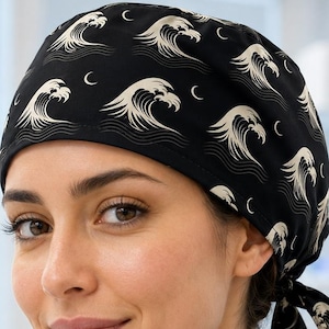 May include: A black scrub cap featuring a repeating pattern of cream-coloured stylised ocean waves and small crescent moons. The cap has a fitted crown and two fabric ties at the back that allow for an adjustable fit, securing the head covering in place.