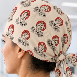 May include: A beige scrub cap featuring a repeating pattern of anatomical hearts centred on red circles with black wave-like details. The cap is secured at the back with two fabric ties, creating a gathered fit around the head for medical use.