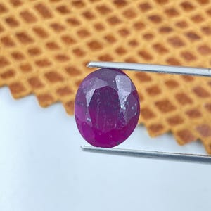 Può includere: Una gemma di rubino sfaccettata, di forma ovale, tenuta da una pinzetta. Il rubino è di un colore rosso-viola intenso e saturo, con inclusioni interne visibili. Lo sfondo è una superficie strutturata di colore marrone chiaro.