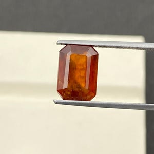 Könnte beinhalten: Ein facettierter, rechteckiger Edelstein, der von einer Pinzette gehalten wird. Der Edelstein hat eine tief orange-rote Farbe mit einer polierten Oberfläche. Die Form des Edelsteins ist ein Smaragdschliff, mit einem rechteckigen Körper und abgeschrägten Ecken. Der Edelstein wird vor einem neutralen Hintergrund gehalten.
