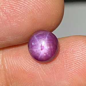 Puede incluir: Una piedra preciosa de rubí estrella de corte cabochon redondo que muestra un tono magenta púrpura profundo. La piedra presenta un efecto de estrella de seis rayos, conocido como asterismo, visible en su superficie pulida y abovedada.