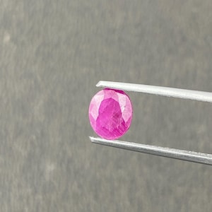 Op de afbeelding: Een gefacetteerde, ovaalvormige robijn edelsteen, vastgehouden door een zilveren pincet. De robijn heeft een levendige roze kleur, met meerdere facetten die het licht vangen. De achtergrond is een effen, neutraal grijs.