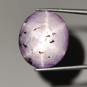 May include: An oval, lavender-coloured gemstone held by tweezers. The gem has a star-like effect with dark inclusions and a polished surface. The stone is set against a neutral grey background.