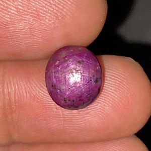 May include: A round, polished, purplish-red gemstone with a star-like pattern. The gem is held between two fingers against a dark background. The stone has a slightly uneven surface with small dark inclusions.