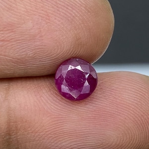 Peut inclure: Une pierre précieuse ronde à facettes d'une couleur rouge magenta profonde et saturée. La pierre présente une taille symétrique avec de multiples facettes triangulaires qui reflètent la lumière sur sa surface, révélant une qualité translucide.