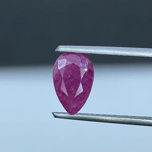 Könnte beinhalten: Ein birnenförmiger, facettierter Edelstein in einem tiefen Magentarosa. Der Stein wird zwischen den beiden Metallspitzen einer Pinzette gehalten und zeigt seine durchscheinende Qualität und den präzisen geometrischen Schliff.