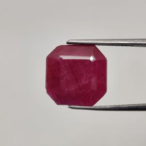 Pode incluir: Uma pedra preciosa de rubi facetada e octogonal, segurada por uma pinça. O rubi é de uma cor vermelha profunda e saturada, com estruturas internas visíveis. A pedra preciosa é cortada com múltiplas facetas para realçar o seu brilho.