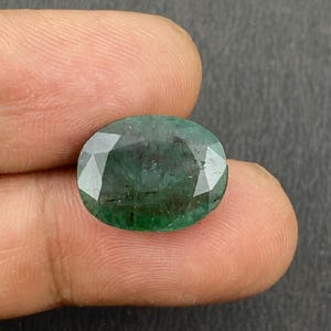 Puede incluir: Una piedra preciosa de esmeralda ovalada y facetada. La gema es de un color verde intenso y rico con inclusiones internas visibles. Las facetas captan la luz, creando un efecto brillante. La piedra preciosa se sostiene entre los dedos.