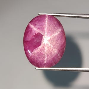 Pode incluir: Gema de rubi oval polida, segurada por pinças. A gema exibe um efeito de estrela de seis raios, com um tom rosa profundo e um padrão em forma de estrela mais claro em sua superfície. A pedra é lisa e reflexiva.