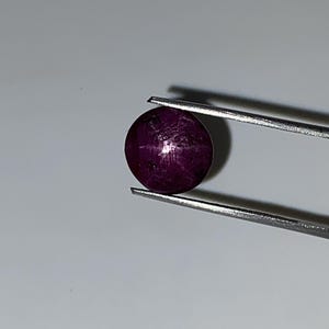 May include: A round, deep red gemstone, held by silver tweezers. The gem has a polished surface with a star-like effect. The background is a plain, light gray. This gemstone is likely a ruby.