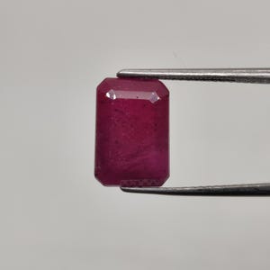 May include: A rectangular, faceted ruby gemstone held by tweezers. The ruby is a deep, rich red color with a slight translucence. The gemstone's cut features a series of flat surfaces, creating a sparkling effect. The tweezers are silver-colored.