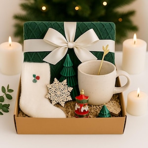 Könnte beinhalten: Eine Weihnachtsgeschenkbox mit einer grünen Strickdecke, die mit einem weißen Band gebunden ist, einer weißen Tasse mit einem goldenen Löffel, einem weißen Strumpf und dekorativen Ornamenten. Die Box enthält Kerzen und einen Weihnachtsbaum im Hintergrund.