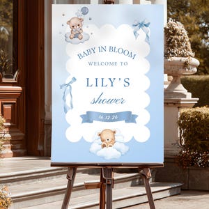Könnte beinhalten: Ein rechteckiges Willkommensschild mit hellblauem Hintergrund und weißem, gewelltem Mittelteil. Es zeigt zwei Teddybären auf Wolken, blaue Schleifen und den Text: BABY IN BLOOM, WELCOME TO, LILY'S shower sowie das Datum 16.12.26 in dunkelblauer Schrift.