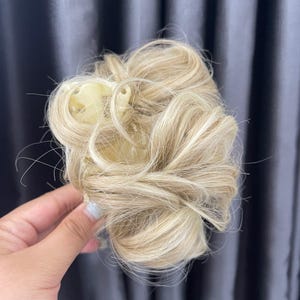 May include: A blonde hair extension piece styled in a messy bun, featuring light golden and platinum highlights. The hair is attached to a cream-colored plastic claw clip, designed to secure the synthetic strands into a voluminous, textured updo.