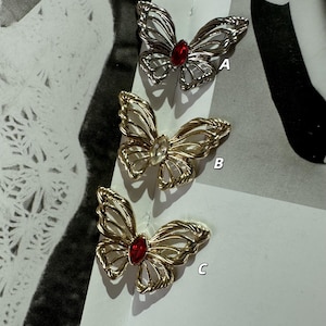 May include: Three butterfly brooches with intricate wireframe wings and jewel centers. One is silver with a red jewel, another gold with a clear jewel, and the third gold with a red jewel. The brooches are displayed on a neutral background.