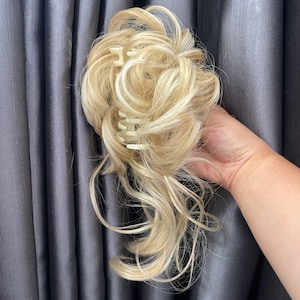 Puede incluir: Un postizo de cabello rubio con mechones sintéticos ondulados y claros dispuestos en un estilo de moño desordenado. El cabello está sujeto a una pinza de plástico de color claro, parcialmente visible a través de los rizos voluminosos.