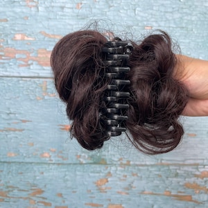 May include: A dark brown hair bun extension attached to a black plastic claw clip. The synthetic hair is styled in a messy, voluminous texture with loose strands, secured centrally by the interlocking teeth of the dark, durable hair accessory.