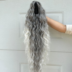 May include: A long, wavy gray and white hair extension with a black clip. The hairpiece is held up against a white door. The hair has a natural, textured appearance, with a blend of gray and white strands.
