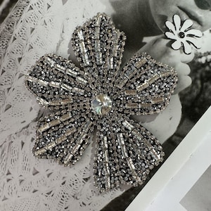 May include: A large, silver-toned flower brooch with a central clear crystal and a petal design encrusted with small, rectangular rhinestones. The brooch is set against a blurred background with a small, white and black flower.