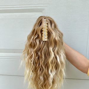 以下が含まれることがあります： 金色のヘアクリップが付いた、ウェーブのかかったブロンドのヘアピース。 ヘアピースは、明るいブロンドと暗いブロンドのトーンが混ざり合い、自然な外観を作り出しています。 クリップは上部に配置され、髪をまとめています。