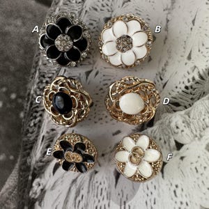 May include: A collection of six decorative buttons with floral designs. The buttons feature a variety of colors, including black, white, and gold, and are embellished with rhinestones. The buttons are round and have a vintage aesthetic.