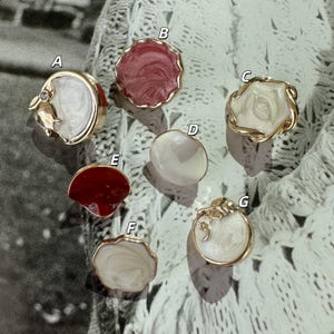 May include: A collection of seven gold-toned, decorative buttons with various designs. The buttons feature white, red, and pink enamel in different shapes, including a flower, a seashell, and a scalloped edge. Some have small embellishments.