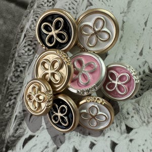 Op de afbeelding: Een verzameling decoratieve knopen in verschillende kleuren, waaronder zwart, wit, roze en goud. Elke knoop heeft een bloemmotief in het midden en een goudkleurige rand. Op sommige knopen staat de inscriptie "Winner" aan de zijkant.