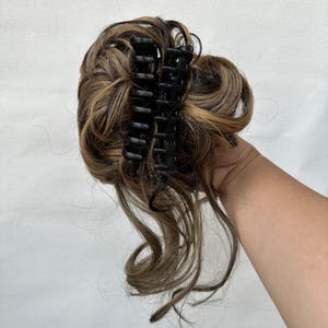 May include: A hair extension piece featuring wavy, multi-tonal brown and blonde synthetic hair strands gathered into a messy bun. The hair is secured with a black plastic claw clip that has interlocking teeth to hold the style in place.