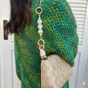 Peut inclure: Un sac à main beige avec une chaîne dorée et des perles. Le sac présente un motif répétitif et une fermeture éclair. La chaîne est fixée avec des anneaux dorés. L'arrière-plan est un pull vert.