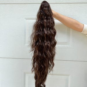 Op de afbeelding: Een donkerbruine, golvende haarextensie, vastgehouden voor een witte deur. Het haarstuk is lang en heeft een natuurlijke, getextureerde uitstraling, met losse krullen en een lichte glans. Het haar heeft een rijke, warme bruine kleur.