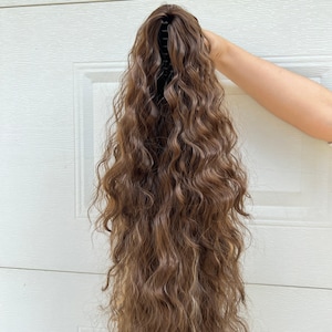 May include: A long, wavy brown hair extension with a black claw clip. The hair has highlights and a natural look. The extension is held up against a white background.