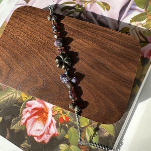 May include: A beaded bracelet featuring a mix of colorful beads, including red, purple, and iridescent beads, along with a metallic flower charm. The bracelet is displayed on a wooden surface, showcasing its intricate design and craftsmanship.