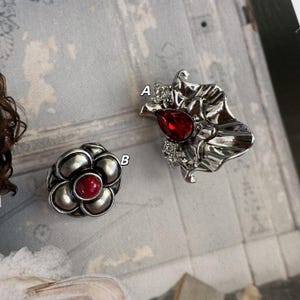 Op de afbeelding: Twee zilverkleurige broches. Eén heeft de vorm van een bloem met een rode kern. De andere is een sierlijk ontwerp met een rode druppelvormige steen en heldere accenten. Beide zijn waarschijnlijk sieraden.